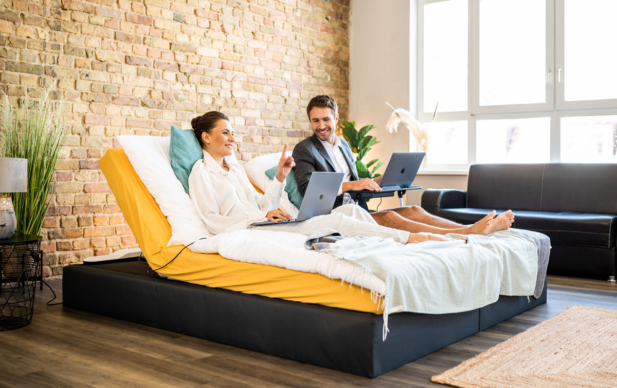 Woman and man sit in box spring bed with CosyLift angled and work on laptop
